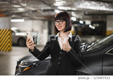 Young Caucasian businesswoman in car park holding smartphone, smiling and giving thumbs up during video call. Professional attire, engaging in conversation, technology focused and modern setting. Young Caucasian businesswoman in car park holding smartphone, smiling and giving thumbs up during video call. Professional attire, engaging in conversation, technology focused and modern setting. 123067422