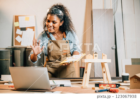 Carpenter woman in a workshop holding furniture design sketches, presenting her project to a customer using a laptop, surrounded by unfinished wooden pieces and tools, craftsmanship and creativity 123067938