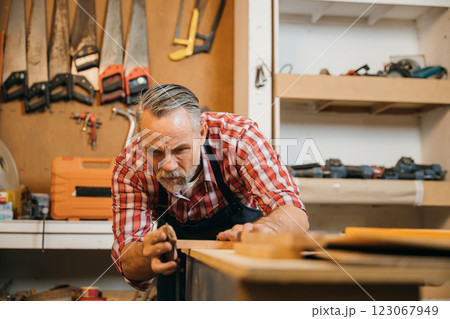 A senior carpenter in red plaid shirt carefully sands piece of wood in his workshop. Tools hang organized on wall behind him, illustrating his skill, dedication and attention to detail in woodworking 123067949