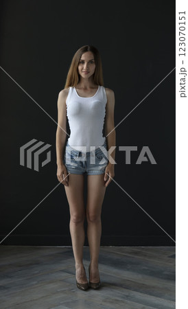 young woman in a white tank top and denim shorts on a dark background, full length portrait 123070151