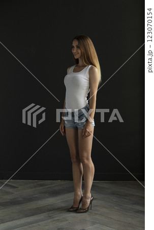 young woman in a white tank top and denim shorts on a dark background, full length portrait 123070154