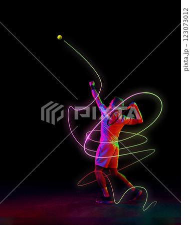 Focused man, tennis player in motion, serving ball with racket with neon green and pink light trails emphasizing motion against black background. Focused man, tennis player in motion, serving ball with racket with neon green and pink light trails emphasizing motion against black background. 123073012