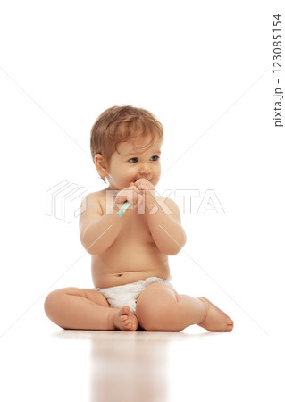 Little baby sitting, wearing in diaper, cross-legged while holding toothbrush and chewing on it against white background. 123085154