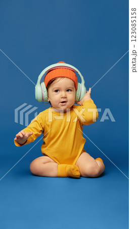 Happy baby wearing bright orange beanie and headphones adjusting ear cups while playfully and funny dancing against deep blue background. Happy baby wearing bright orange beanie and headphones adjusting ear cups while playfully and funny dancing against deep blue background. 123085158