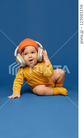 Smiling baby wearing bright orange beanie and oversized headphones sitting on blue floor against deep blue background Smiling baby wearing bright orange beanie and oversized headphones sitting on blue floor against deep blue background 123085159
