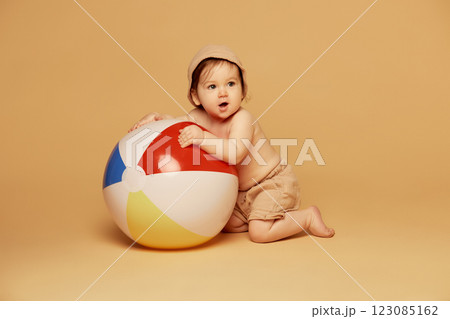 Portrait of playful little baby, wearing beige shorts and hat, hugging large colorful beach ball while kneeling against warm beige background. 123085162