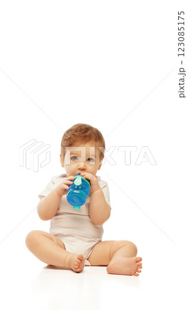 Little baby, dressed white onesie, sitting and drinking from blue sippy cup while holding it with both hands against white background. 123085175