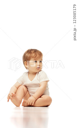 Baby, dressed white onesie, sitting cross-legged while looking sideways with focused expression against white background. Baby, dressed white onesie, sitting cross-legged while looking sideways with focused expression against white background. 123085179