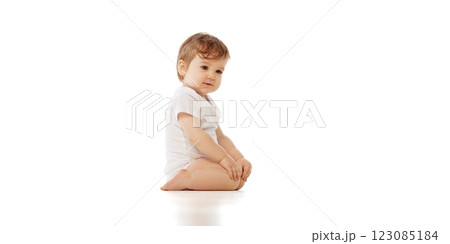 Little baby, dressed white onesie, sitting with hands on knees while looking slightly downward against white background. 123085184