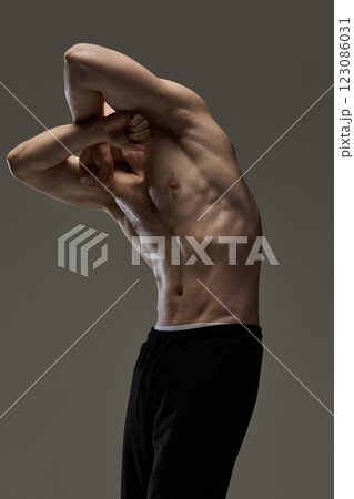 Man dressed black, stylish pants, twisting arms in artistic pose with flexed abs under dramatic lighting against deep brown background. 123086031