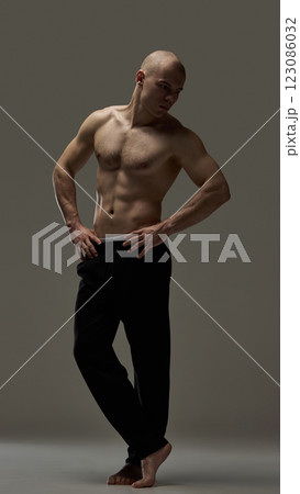 Full length photo of shirtless athletic man in black pants standing in powerful posture with sculpted abs against moody background. 123086032