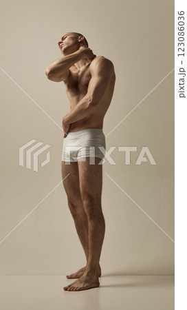 Shirtless male model dressed white underwear, tilting head with hand on neck in expressive movement against minimal studio background. 123086036