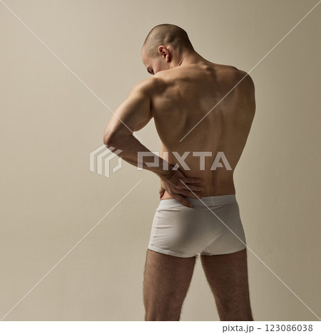 Back view of young, strong man holding lower back in poised stance with sculpted muscles against clean background. 123086038