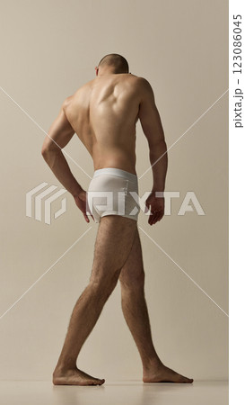 Shirtless man, dressed underwear walking forward showing powerful back muscles emphasized by soft lighting against beige studio background. 123086045