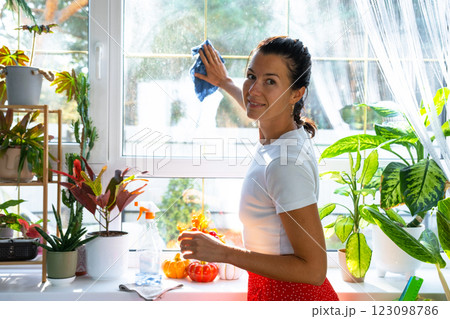 Woman manually washes the window of house with rag cleaner and mop inside the interior with home plants on windowsill and pumpkin decor. Restoring order and cleanliness in autumn, cleaning servise 123098786