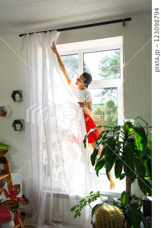 Woman hangs transparent tulle curtains on large windows in the house inside the interior with potted plants. Spring cleaning, tidying up 123098794