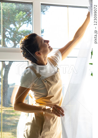 Woman in apron hangs transparent tulle curtains on large windows in the house inside the interior with potted plants. Spring cleaning, tidying up 123099072