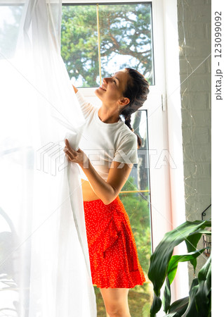 Woman hangs transparent tulle curtains on large windows in the house inside the interior with potted plants. Spring cleaning, tidying up Woman hangs transparent tulle curtains on large windows in the house inside the interior with potted plants. Spring cleaning, tidying up 123099092