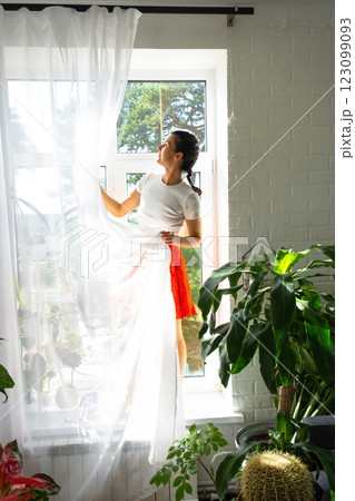 Woman hangs transparent tulle curtains on large windows in the house inside the interior with potted plants. Spring cleaning, tidying up Woman hangs transparent tulle curtains on large windows in the house inside the interior with potted plants. Spring cleaning, tidying up 123099093