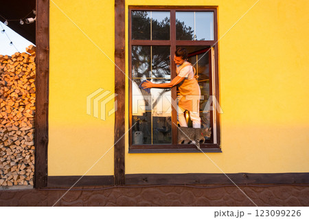 Woman in apron manually washes the window of the house with a rag with a spray cleaner and a mop outside. Safety at height, restoring order and cleanliness in the spring, cleaning service Woman in apron manually washes the window of the house with a rag with a spray cleaner and a mop outside. Safety at height, restoring order and cleanliness in the spring, cleaning service 123099226
