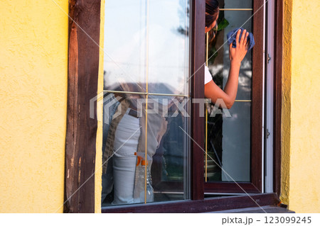 Woman in apron manually washes the window of the house with a rag with a spray cleaner and a mop outside. Safety at height, restoring order and cleanliness in the spring, cleaning service 123099245