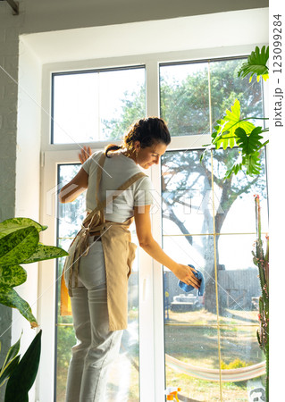 Woman in apron manually washes the window of house with rag cleaner and mop inside the interior with home plants on windowsill. Restoring order and cleanliness in the spring, cleaning servise Woman in apron manually washes the window of house with rag cleaner and mop inside the interior with home plants on windowsill. Restoring order and cleanliness in the spring, cleaning servise 123099284