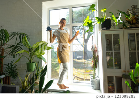 Woman Portrait in apron manually washes the window of house with rag cleaner and mop inside the interior with home plants on windowsill. Restoring order and cleanliness in the spring, cleaning servise Woman Portrait in apron manually washes the window of house with rag cleaner and mop inside the interior with home plants on windowsill. Restoring order and cleanliness in the spring, cleaning servise 123099285