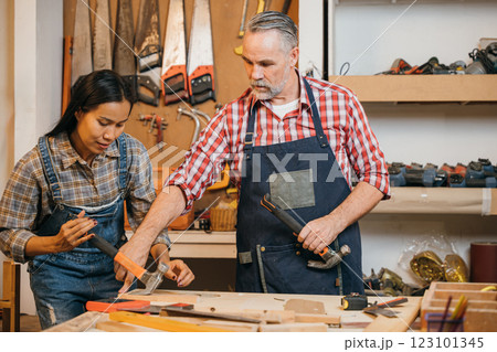 Senior carpenter teaches a woman woodworking techniques, guiding her as she uses a hammer on a wood project. They work together at a well-equipped workbench in a detailed, organized workshop Senior carpenter teaches a woman woodworking techniques, guiding her as she uses a hammer on a wood project. They work together at a well-equipped workbench in a detailed, organized workshop 123101345