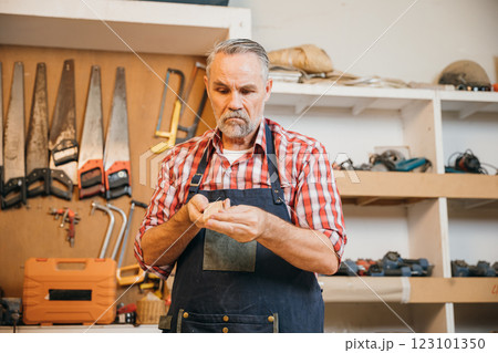 A senior carpenter in red plaid shirt carefully sands piece of wood in his workshop. Tools hang organized on wall behind him, illustrating his skill, dedication and attention to detail in woodworking 123101350