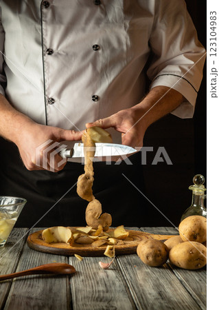 A talented chef demonstrates culinary skill while peeling a potato with a sharp knife. Stunning golden peels spiral down onto a wooden board surrounded by fresh ingredients 123104983