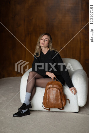 A girl in a warm sweater, a black short skirt, nylon tights, and black shoes is posing with a brown backpack. 123107191