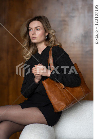 A girl in a warm sweater, a black short skirt, nylon tights, and black shoes is posing with a brown bag. 123107195