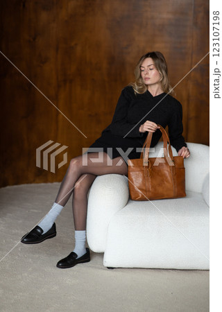 A girl in a warm sweater, a black short skirt, nylon tights, and black shoes is posing with a brown bag. 123107198