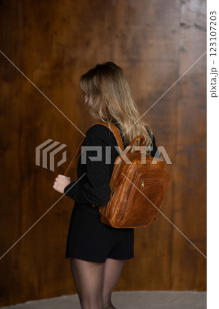 A girl in a warm sweater, a black short skirt and nylon tights is posing with a brown backpack. 123107203