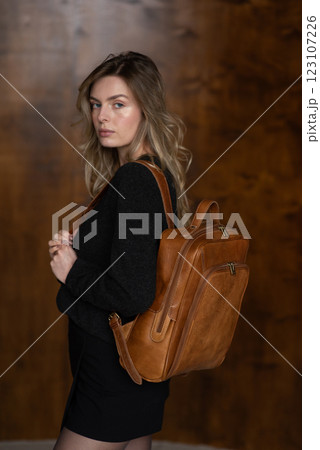 A girl in a warm sweater, a black short skirt and nylon tights is posing with a brown backpack. 123107226