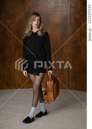 A girl in a warm sweater, a black short skirt, nylon tights, and black shoes is posing with a brown backpack. 123107243