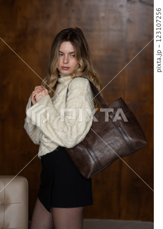A girl in a warm sweater, a black short skirt, nylon tights is posing with a brown leather bag. 123107256