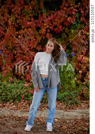 Stylish young woman in trendy autumn outfit posing confidently in a park with vibrant fall foliage, wearing a black-and-white plaid shirt over a white crop top, paired with blue jeans and white Stylish young woman in trendy autumn outfit posing confidently in a park with vibrant fall foliage, wearing a black-and-white plaid shirt over a white crop top, paired with blue jeans and white 123110587