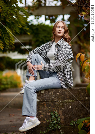 Stylish young woman in casual autumn outfit sitting on a stone wall in a green park with orange leaves, wearing a black and white plaid shirt, jeans, and sneakers – outdoor fashion portrait Stylish young woman in casual autumn outfit sitting on a stone wall in a green park with orange leaves, wearing a black and white plaid shirt, jeans, and sneakers – outdoor fashion portrait 123110591