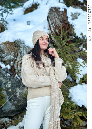 Young woman in cream-colored knitted sweater and beanie standing in a serene snowy mountain landscape with rocky outcrops and evergreen trees, exuding a contemplative and wintery atmosphere. Young woman in cream-colored knitted sweater and beanie standing in a serene snowy mountain landscape with rocky outcrops and evergreen trees, exuding a contemplative and wintery atmosphere. 123110599
