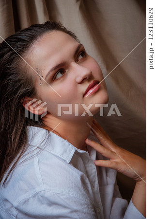 Thoughtful young woman with gemstones on face in relaxed pose. Caucasian woman with glitter on face. Beige background 123110629