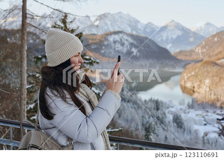 Winter Wanderlust: Young Woman Using Smartphone in Snowy Mountain Landscape with Golden Sunlight and Picturesque Village 123110691