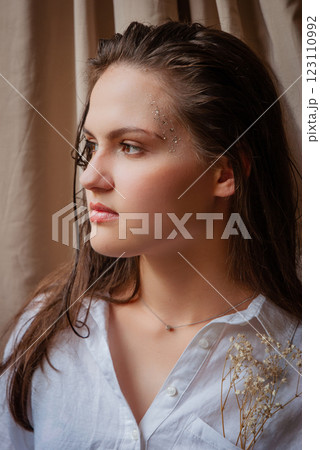 Thoughtful woman in white shirt with glitter makeup and dried flowers. Caucasian woman with glitter on face. Beige background 123110992