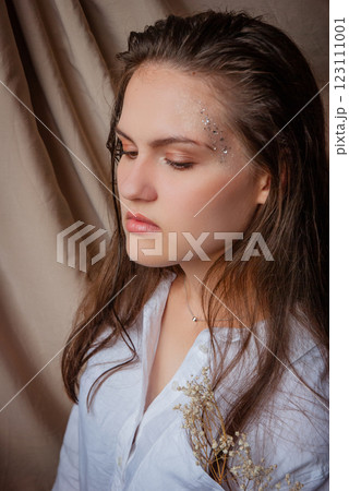 Reflective woman with sparkles and white flowers against a draped fabric background. Caucasian woman with glitter on face. Beige background 123111001