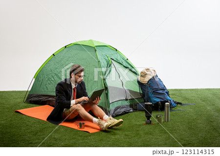 Businessman in shorts sitting on camping mat using laptop, holding coffee cup against white studio background. Businessman in shorts sitting on camping mat using laptop, holding coffee cup against white studio background. 123114315