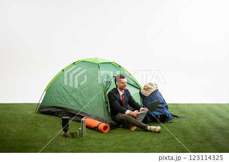 Businessman sitting near camping tent working on laptop, smiling, leaning on backpack against white studio background. 123114325