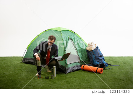 Businessman crouching near camping tent lifting coffee press while working on laptop against white studio background. Businessman crouching near camping tent lifting coffee press while working on laptop against white studio background. 123114326