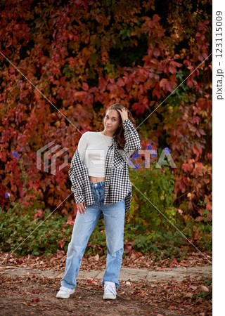 Stylish young woman in trendy autumn outfit posing confidently in a park with vibrant fall foliage, wearing a black-and-white plaid shirt over a white crop top, paired with blue jeans and white Stylish young woman in trendy autumn outfit posing confidently in a park with vibrant fall foliage, wearing a black-and-white plaid shirt over a white crop top, paired with blue jeans and white 123115009
