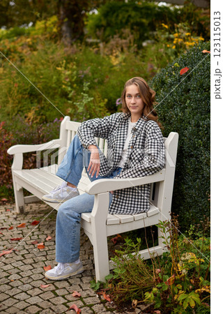 Young Women Sitting on a White Wooden Bench in a Tranquil Garden with Lush Greenery and Casual Attire 123115013