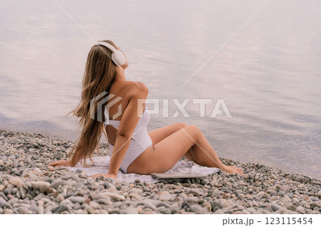 A woman is sitting on a beach with her headphones on. She is wearing a white bikini and a white shirt. The beach is rocky and the water is calm. The woman is enjoying her time by the water. A woman is sitting on a beach with her headphones on. She is wearing a white bikini and a white shirt. The beach is rocky and the water is calm. The woman is enjoying her time by the water. 123115454